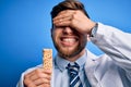 Young blond doctor man with beard and blue eyes wearing coat eating granola bar stressed with hand on head, shocked with shame and Royalty Free Stock Photo