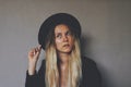 Young blond female wiccan witch wearing a black witch hat and black blazer. Artsy portrait against dark grey wall