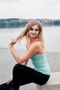 Young blond fit slim woman, sitting in front of city lake in summer. Three-quarter picture of fitness trainer, wearing black