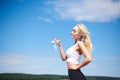 Young blond fit woman, wearing black leggings and white top,drinking water from glass bottle outside with blue sunny sky on Royalty Free Stock Photo