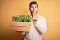 Young blond gardener man with beard and blue eyes holding wooden box with plants cover mouth with hand shocked with shame for