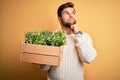 Young blond gardener man with beard and blue eyes holding wooden box with plants serious face thinking about question, very