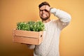 Young blond gardener man with beard and blue eyes holding wooden box with plants stressed with hand on head, shocked with shame