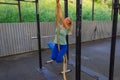 Young blond girl in blue leggings and green t-shirt smiling climbing up the rope in the gym among the horizontal bars against Royalty Free Stock Photo