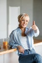 A young blond girl in stylish glasses sits in the kitchen with a glass of juice. Looks at the camera and shows the super gesture Royalty Free Stock Photo