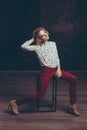 A young blond handsome man androgyn in the image of an attractive woman posing sitting on a chair on a dark background