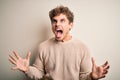 Young blond handsome man with curly hair wearing casual sweater over white background crazy and mad shouting and yelling with Royalty Free Stock Photo