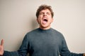 Young blond handsome man with curly hair wearing casual sweater over white background crazy and mad shouting and yelling with Royalty Free Stock Photo