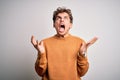 Young blond handsome man with curly hair wearing casual sweater over white background crazy and mad shouting and yelling with Royalty Free Stock Photo