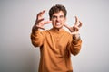 Young blond handsome man with curly hair wearing casual sweater over white background Shouting frustrated with rage, hands trying Royalty Free Stock Photo