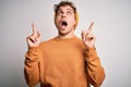 Young blond handsome man with curly hair wearing casual sweater and wool hat amazed and surprised looking up and pointing with Royalty Free Stock Photo