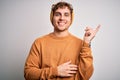 Young blond handsome man with curly hair wearing casual sweater and wool hat with a big smile on face, pointing with hand and Royalty Free Stock Photo