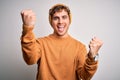 Young blond handsome man with curly hair wearing casual sweater and wool hat celebrating surprised and amazed for success with Royalty Free Stock Photo