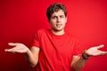 Young blond handsome man with curly hair wearing casual t-shirt over red background clueless and confused with open arms, no idea Royalty Free Stock Photo