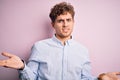 Young blond handsome man with curly hair wearing striped shirt over white background clueless and confused with open arms, no idea Royalty Free Stock Photo