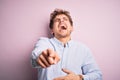 Young blond handsome man with curly hair wearing striped shirt over white background laughing at you, pointing finger to the Royalty Free Stock Photo