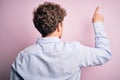 Young blond handsome man with curly hair wearing striped shirt over white background Posing backwards pointing ahead with finger