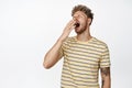 Young blond handsome man yawning from fatigue, standing sleepy, cover yawn with hand near opened mouth, standing over