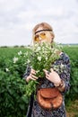 Young blond hippie woman, wearing grey boho style dress and yellow sunglasses, standing on green field, holding camomile bouquet, Royalty Free Stock Photo