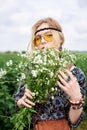 Young blond hippie woman, wearing grey boho style dress and yellow sunglasses, standing on green field, holding camomile bouquet, Royalty Free Stock Photo