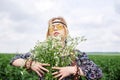 Young blond hippie woman, wearing grey boho style dress and yellow sunglasses, standing on green field, holding camomile bouquet, Royalty Free Stock Photo