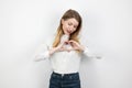Young blond lovely woman making heart symbol with her hands standing on isolated white background, looking gentle,body language,