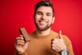 Young blond man with beard and blue eyes eating healthy chocolate protein bar happy with big smile doing ok sign, thumb up with Royalty Free Stock Photo