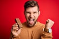 Young blond man with beard and blue eyes eating healthy chocolate protein bar screaming proud and celebrating victory and success Royalty Free Stock Photo