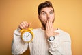 Young blond man with beard and blue eyes holding alarm clock over yellow background cover mouth with hand shocked with shame for Royalty Free Stock Photo