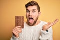 Young blond man with beard and blue eyes holding chocolate bar over yellow background very happy and excited, winner expression Royalty Free Stock Photo