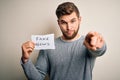 Young blond man with beard and blue eyes holding paper with fake news message pointing with finger to the camera and to you, hand Royalty Free Stock Photo