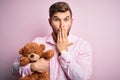 Young blond man with beard and blue eyes holding teddy bear toy over pink background cover mouth with hand shocked with shame for Royalty Free Stock Photo