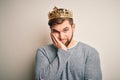 Young blond man with beard and blue eyes wearing golden crown of king thinking looking tired and bored with depression problems
