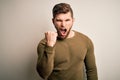 Young blond man with beard and blue eyes wearing green sweater over white background angry and mad raising fist frustrated and Royalty Free Stock Photo