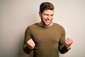Young blond man with beard and blue eyes wearing green sweater over white background very happy and excited doing winner gesture Royalty Free Stock Photo