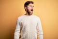 Young blond man with beard and blue eyes wearing white sweater over yellow background angry and mad screaming frustrated and Royalty Free Stock Photo