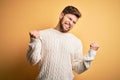 Young blond man with beard and blue eyes wearing white sweater over yellow background very happy and excited doing winner gesture Royalty Free Stock Photo