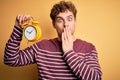 Young blond man with curly hair holding alarm clock standing over isolated yellow background cover mouth with hand shocked with Royalty Free Stock Photo