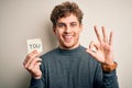 Young blond man with curly hair holding paper with you message over white background doing ok sign with fingers, excellent symbol Royalty Free Stock Photo