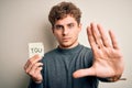 Young blond man with curly hair holding paper with you message over white background with open hand doing stop sign with serious Royalty Free Stock Photo