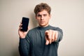 Young blond man with curly hair showing smartphone standing over isolated white background pointing with finger to the camera and Royalty Free Stock Photo