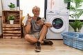 Young blond man doing laundry using smartphone gesturing finger crossed smiling with hope and eyes closed Royalty Free Stock Photo