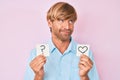 Young blond man holding heart and question mark reminder smiling looking to the side and staring away thinking Royalty Free Stock Photo