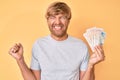 Young blond man holding united kingdom pounds screaming proud, celebrating victory and success very excited with raised arm