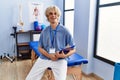 Young blond man pysiotherapist holding document sitting on massage board at rehab clinic