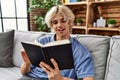 Young blond man reading book sitting on sofa at home Royalty Free Stock Photo