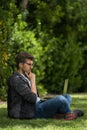 A young blond man is sitting on the grass of a park with his laptop. He is with her legs crossed and he seems to be thinking about Royalty Free Stock Photo