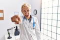 Young blond man wearing doctor uniform and stethoscope at clinic pointing displeased and frustrated to the camera, angry and