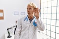 Young blond man wearing doctor uniform and stethoscope at clinic smiling with hand over ear listening an hearing to rumor or Royalty Free Stock Photo