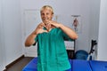 Young blond man wearing physiotherapist uniform standing at clinic smiling in love doing heart symbol shape with hands Royalty Free Stock Photo